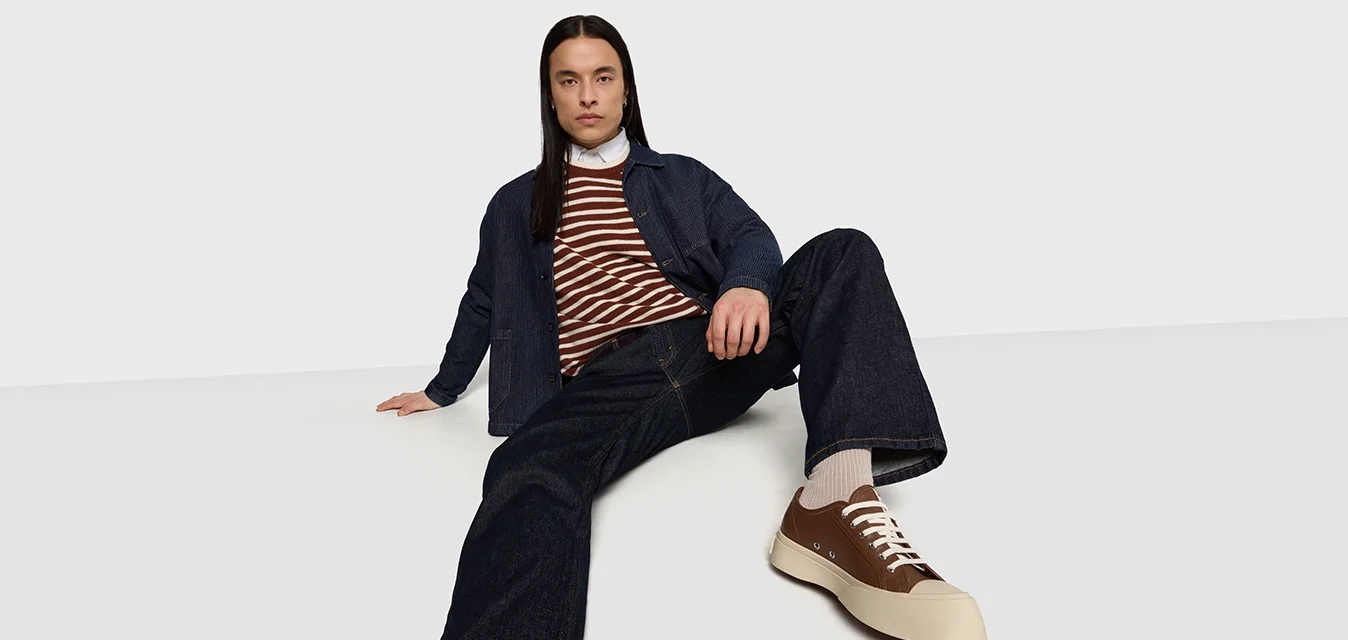 Giubbotto di denim, maglione a righe marroni e bianche, jeans scuri, sneakers beige con accenti marroni, posa seduta, sfondo neutro.
