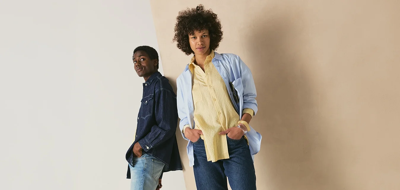 Due persone stanno appoggiate in modo informale a un muro; una indossa una camicia di jeans blu e jeans, l'altra una camicia azzurro chiaro, una maglietta gialla e jeans.