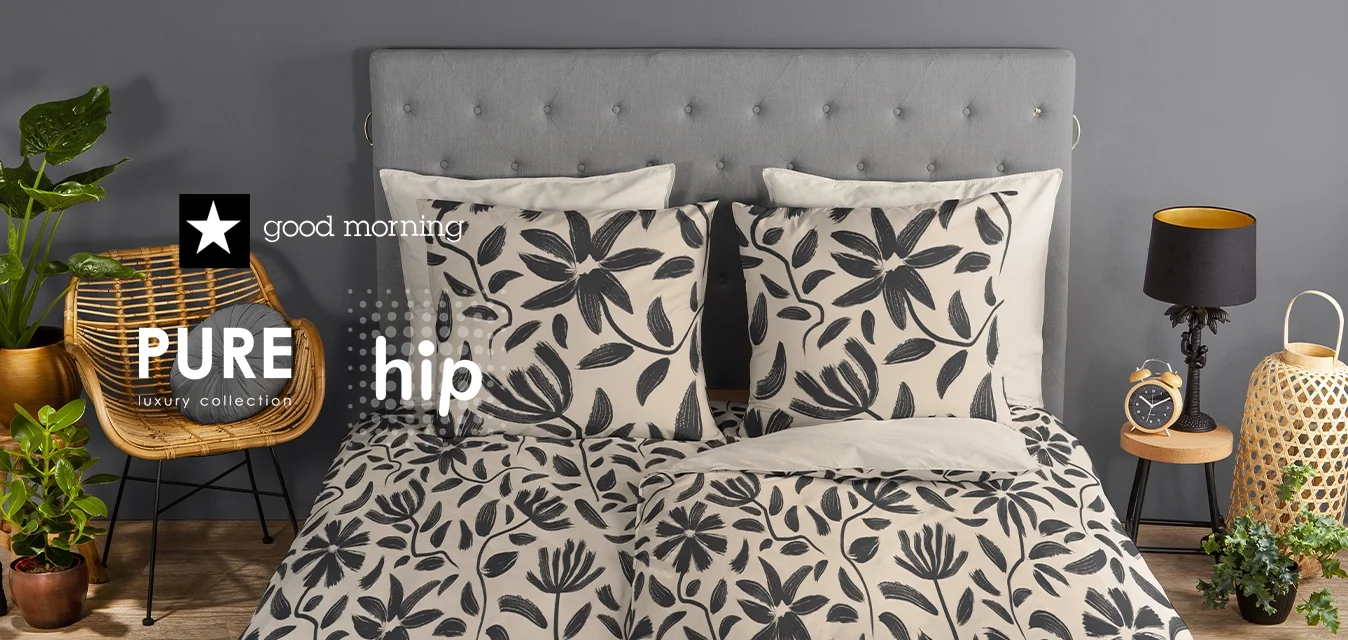 In una camera da letto, un letto rivestito in grigio con un copriletto a motivo floreale in bianco e nero, abbinato a cuscini coordinati. Sedia intrecciata, piante e lampada.
