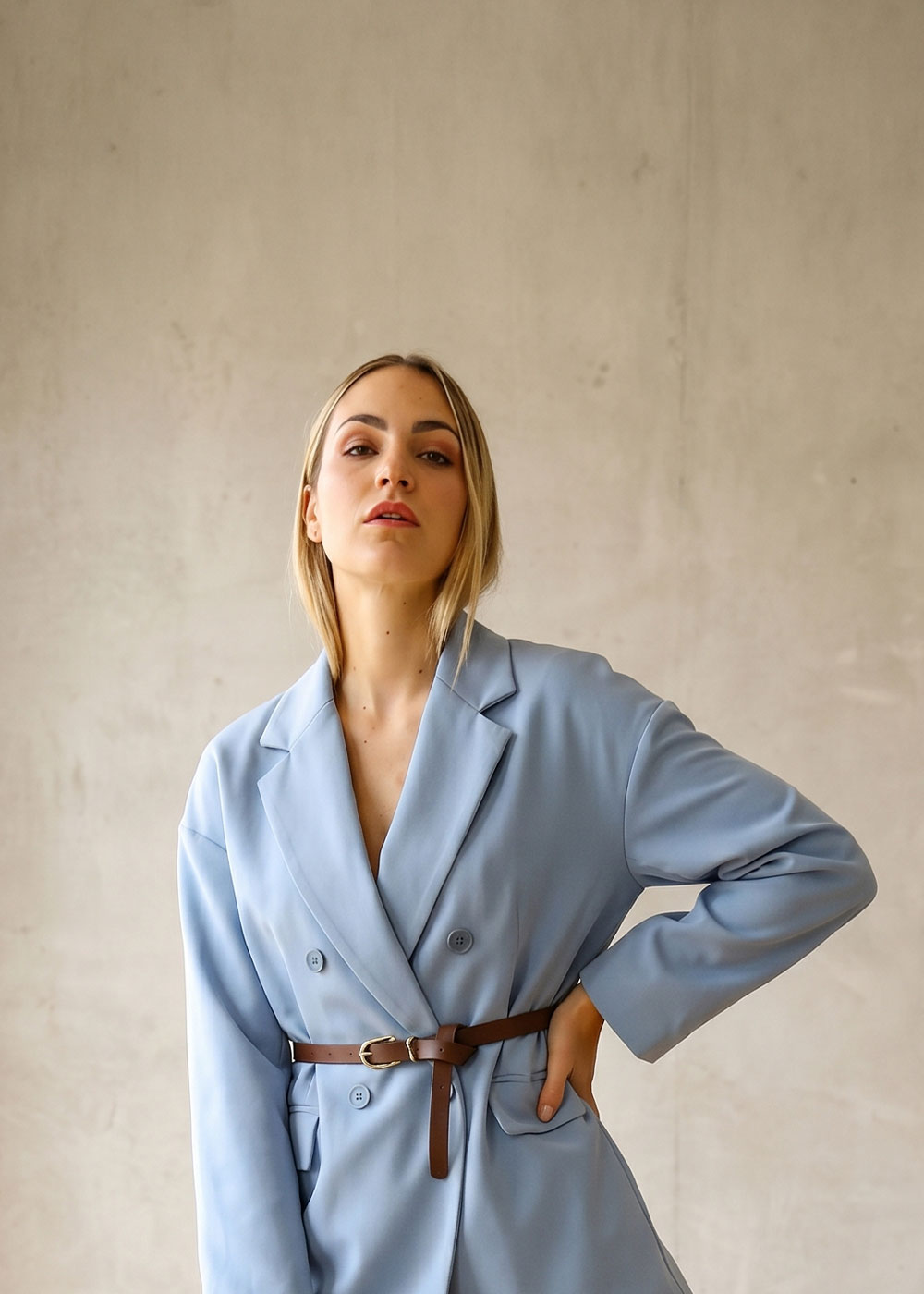 Young woman wearing a light blue double-breasted blazer cinched with a brown belt, hand on hip, posing against a neutral textured backdrop