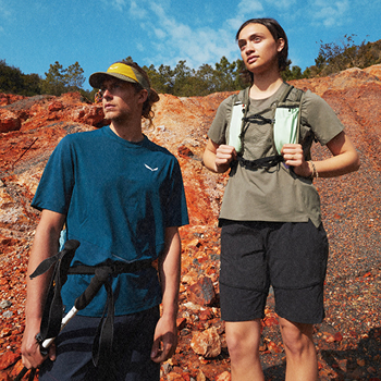 Two hikers (man and woman) in outdoor clothing with backpacks on a red rocky trail under a bright blue sky