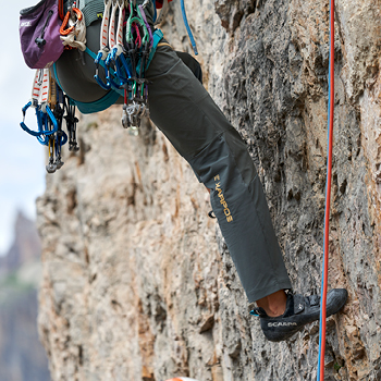 Close-up of a rock climber's legs, harness and carabiners beside a rope on a vertical limestone cliff