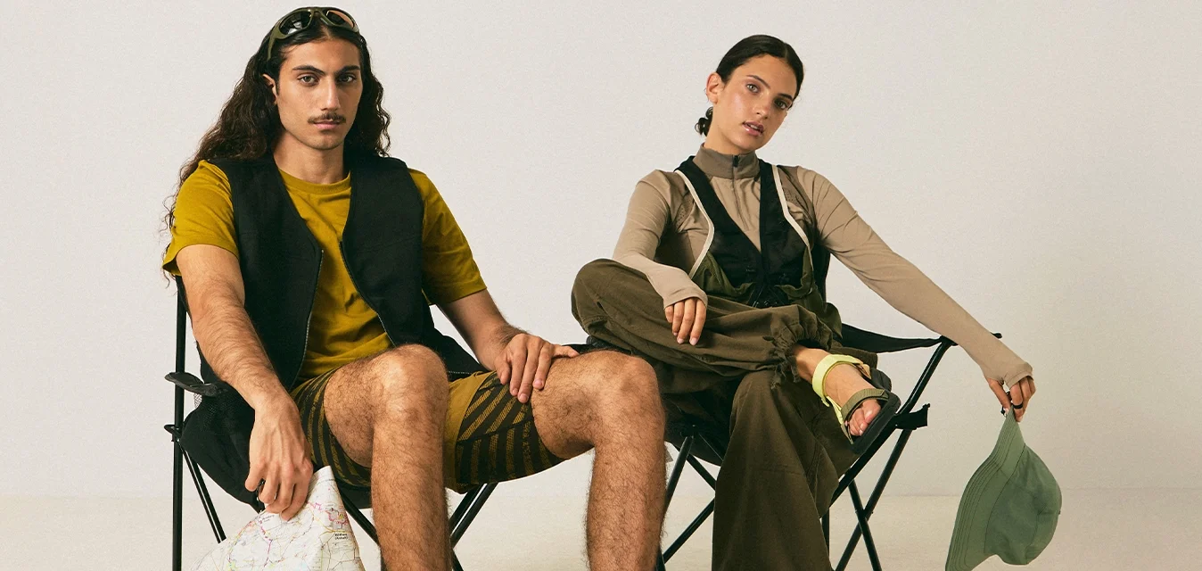 Un uomo e una donna seduti su sedie pieghevoli nere su un pavimento beige, l'uomo tiene una mappa, la donna tiene un cappello verde, entrambi indossano abbigliamento casual.