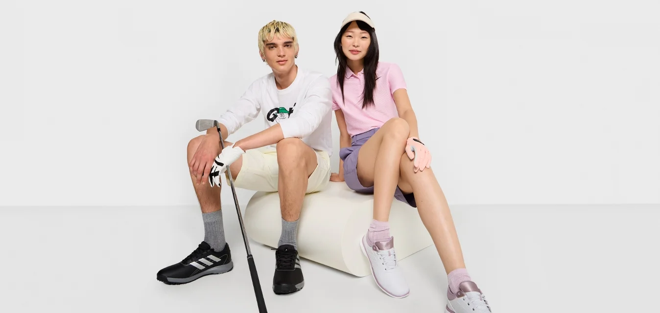 Due persone siedono su una superficie bianca. Uno indossa una maglietta bianca a maniche lunghe con un logo, pantaloni corti beige e scarpe nere; l'altro indossa una polo rosa, pantaloni corti viola e scarpe da ginnastica bianche.