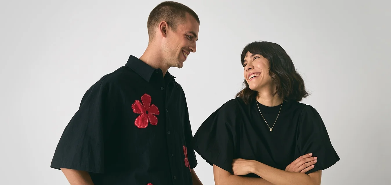 Uomo e donna sorridenti che si guardano, l'uomo indossa una maglietta nera con fiori rossi, la donna indossa un top nero e una gonna bianca su uno sfondo semplice.