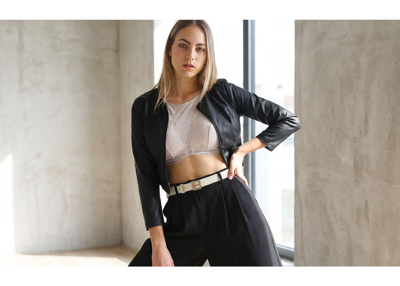Young woman model in black leather jacket and lace crop top, high-waisted trousers with white belt, posing by a window in minimal interior