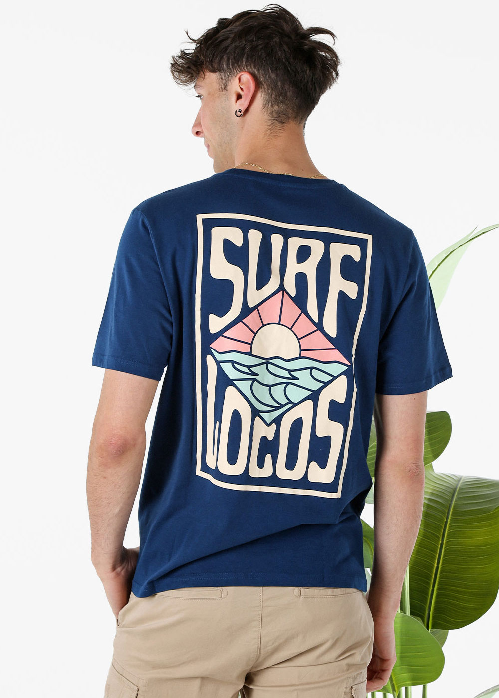 Man seen from behind wearing a navy T-shirt with a large 'SURF LOCOS' sunset and waves graphic, beige shorts and green plant background