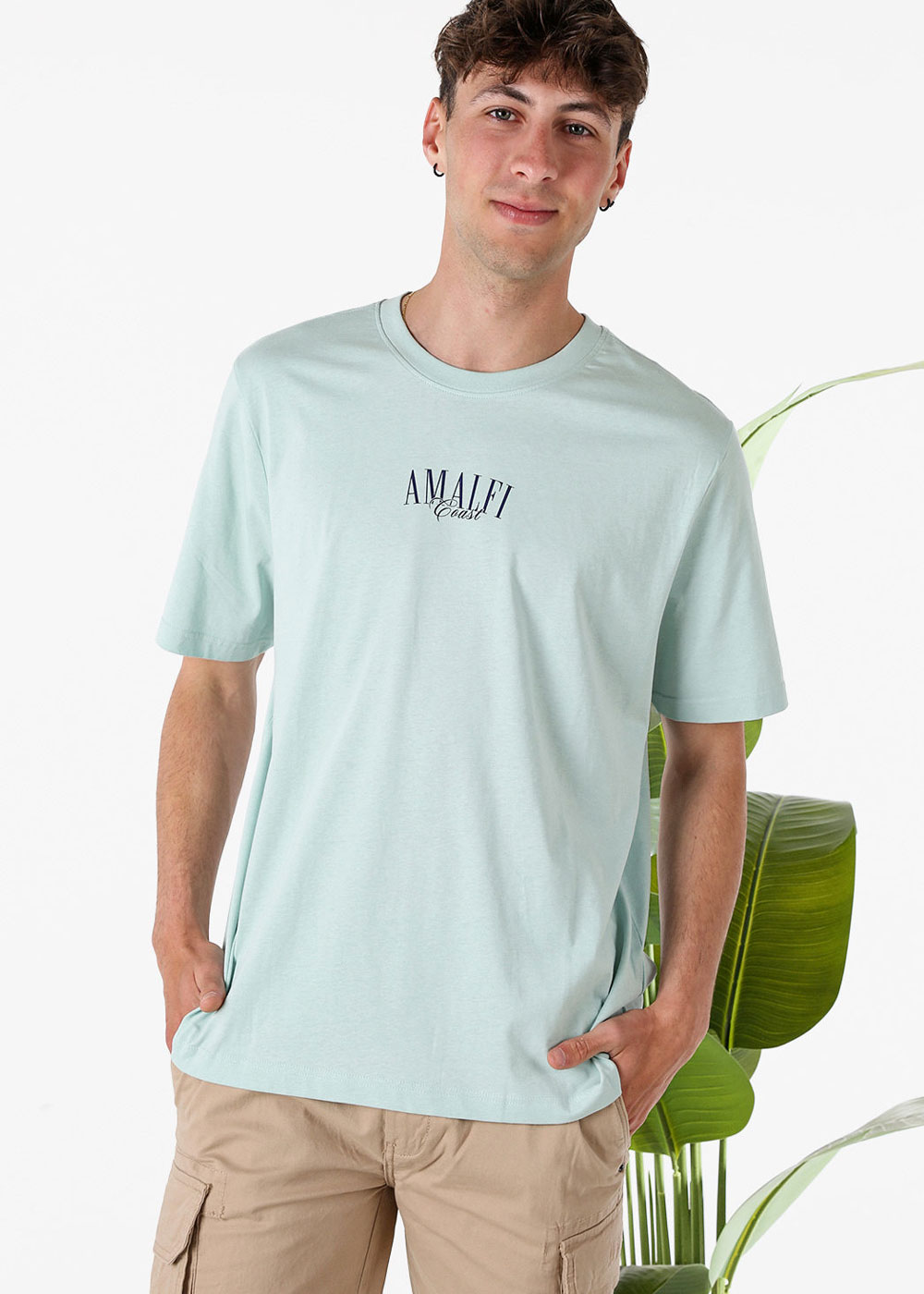 Young man wearing mint t-shirt with "Amalfi Coast" print and khaki cargo shorts, standing by a tropical plant on white background