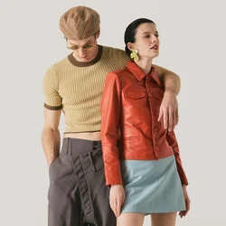 Uomo con cappello marrone pied de poule e camicia corta con pantaloni grigi, braccio intorno a donna con giacca di pelle rossa, gonna blu e stivali beige.