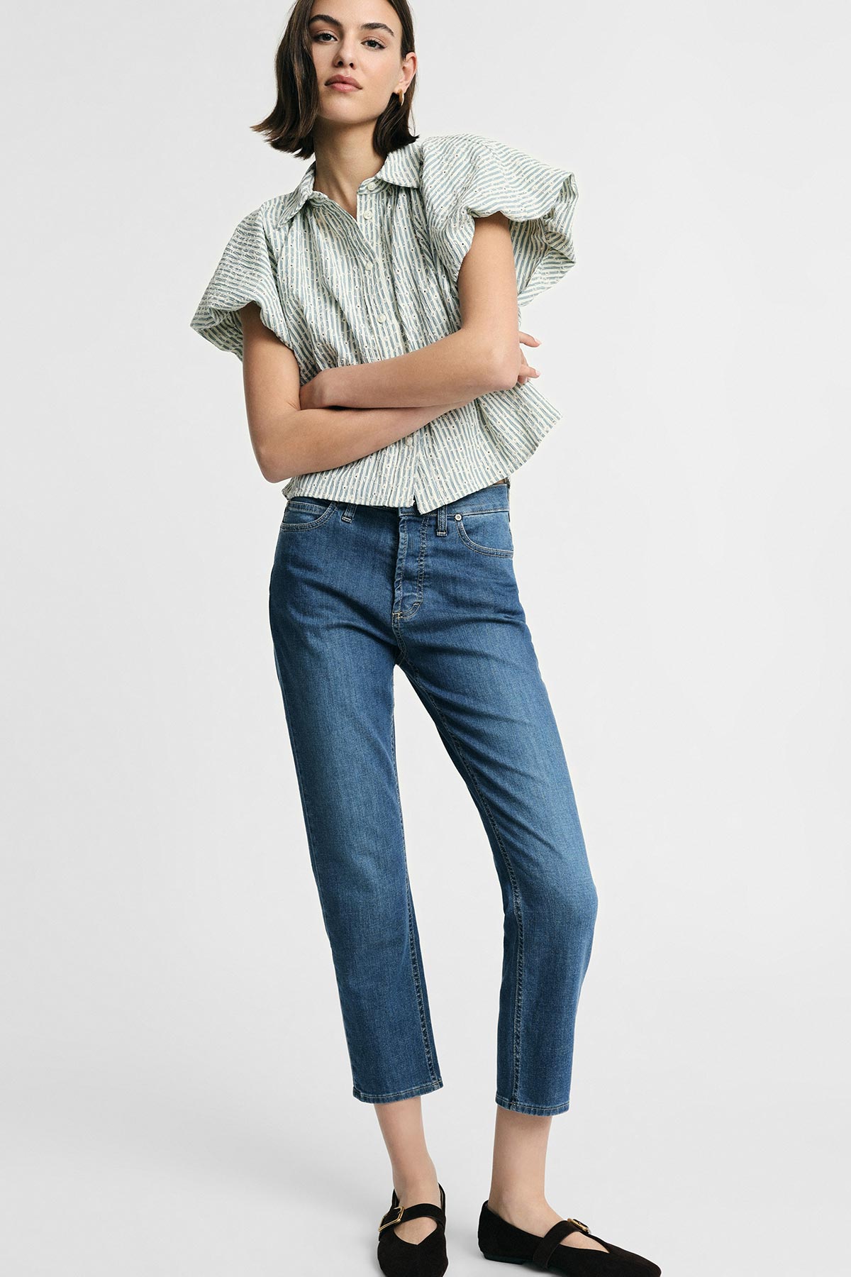 Woman in a striped short-sleeved blouse and cropped blue jeans with black flats standing against a pale background