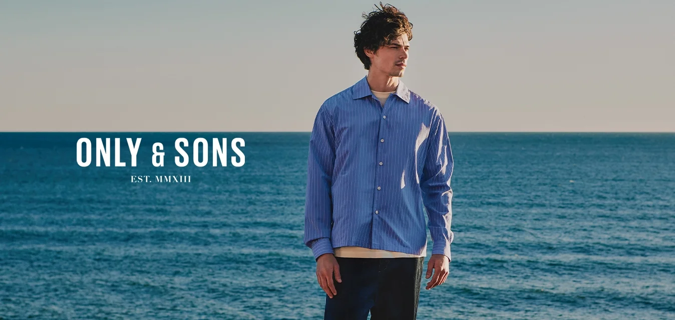 Giovane uomo con una camicia blu che si trova su una costa rocciosa e guarda verso il mare sotto un cielo limpido.