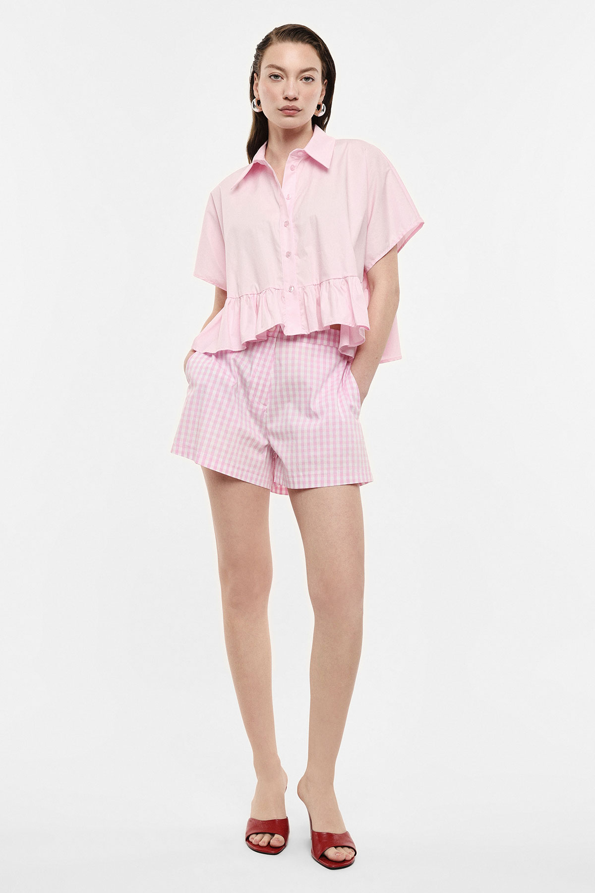 Woman wearing pale pink ruffle-hem short-sleeved shirt and pink gingham shorts with red open-toe heels