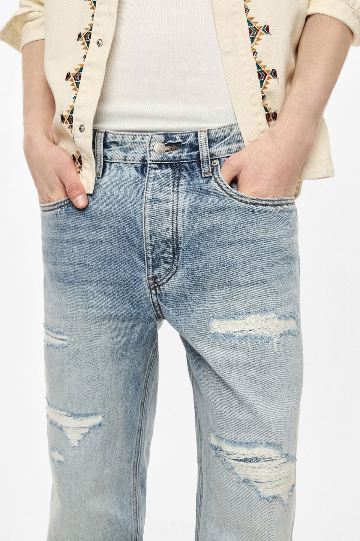 Close-up of midsection wearing light blue ripped jeans with hands in pockets, cream embroidered shirt and white vest