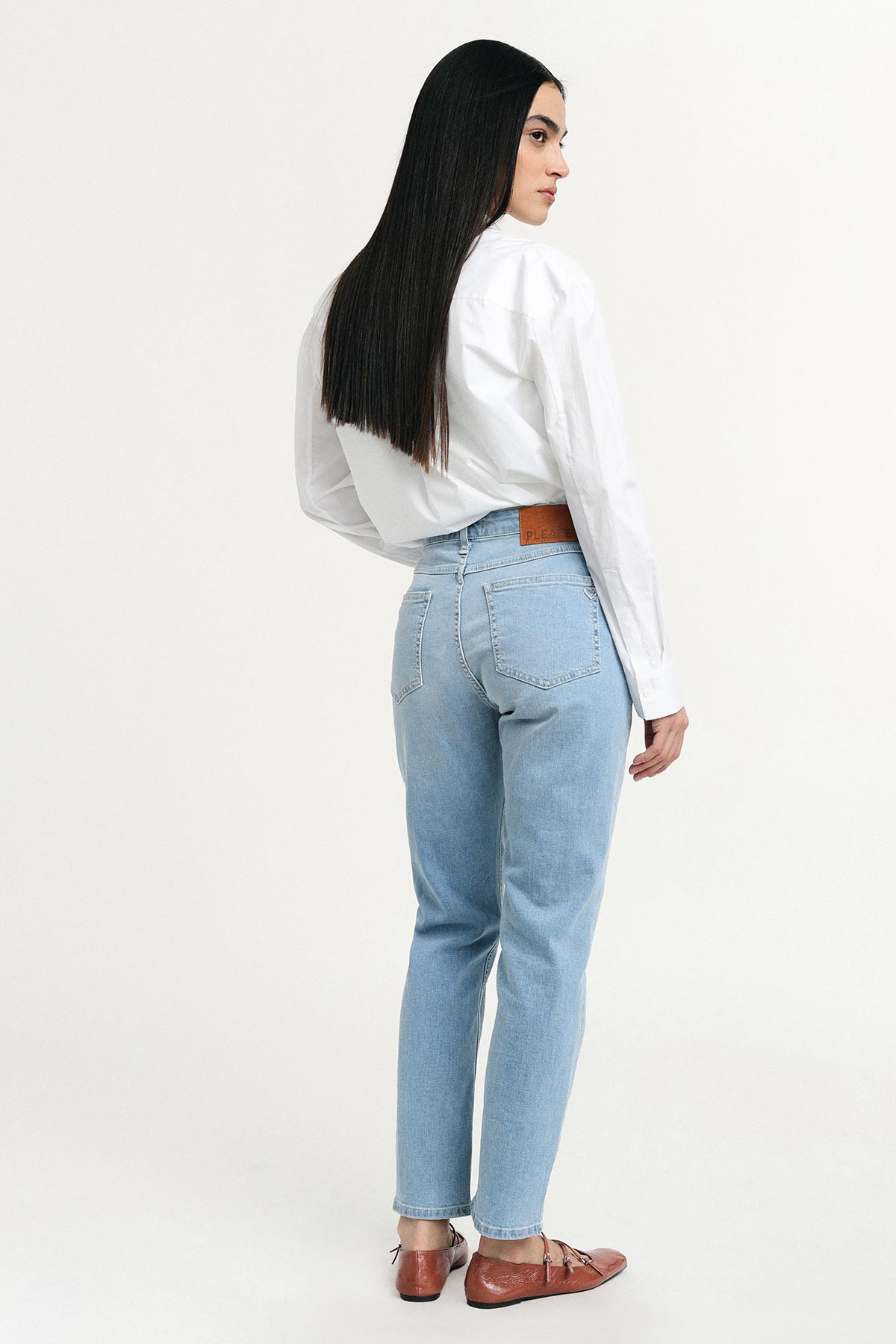 Woman modelling light-blue high-waist jeans and white shirt, brown leather flats, long dark hair, standing looking over shoulder