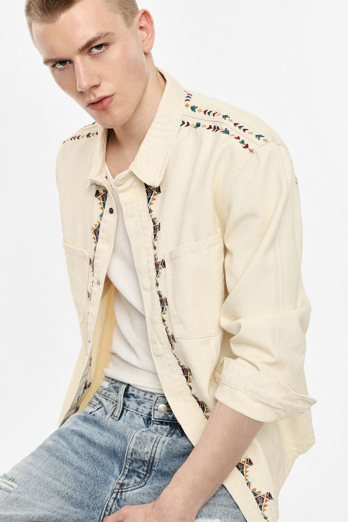 Young man wearing cream embroidered shirt over white vest and faded jeans, seated against plain white background