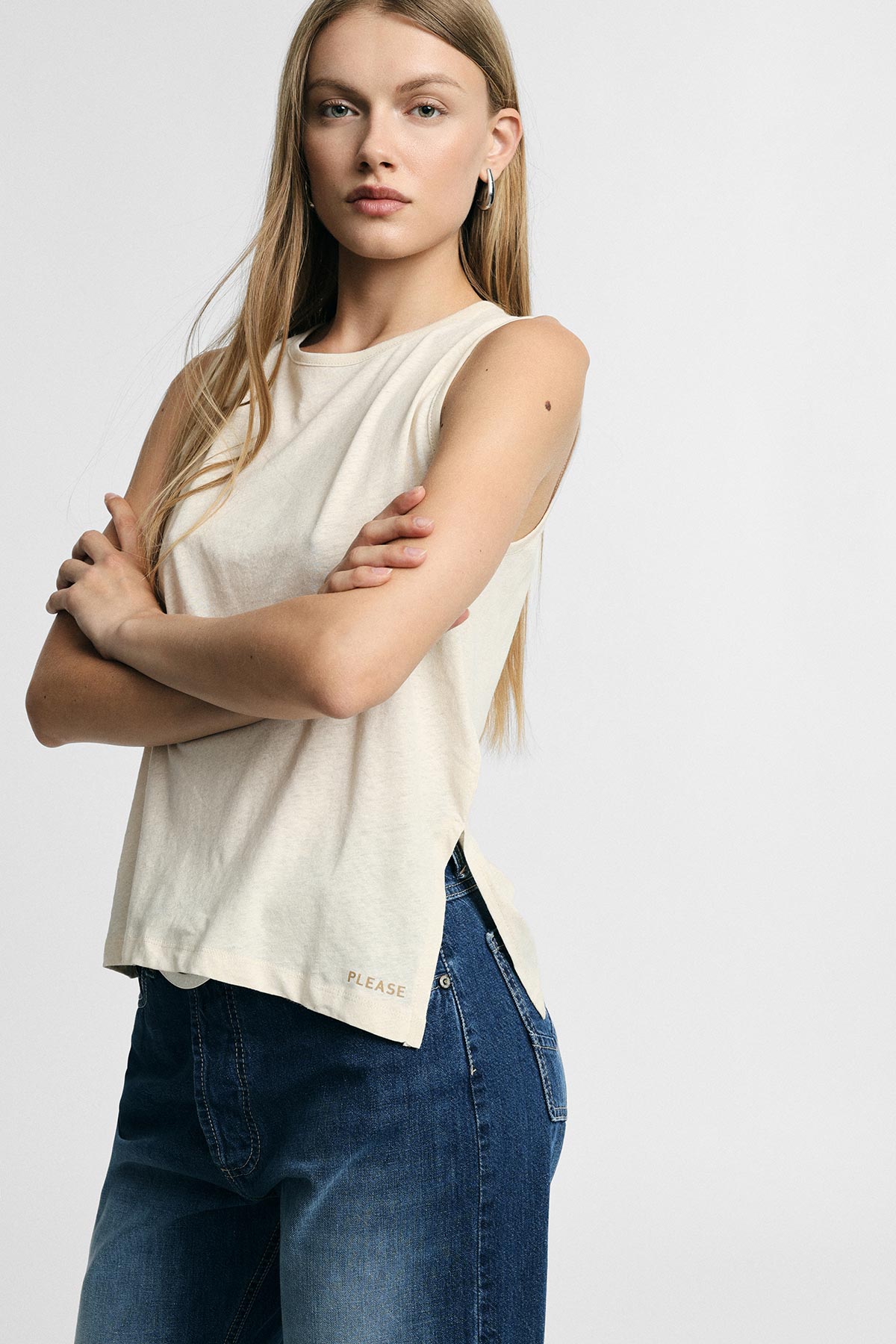 Young woman standing with arms crossed wearing a sleeveless cream top and blue jeans on a plain background