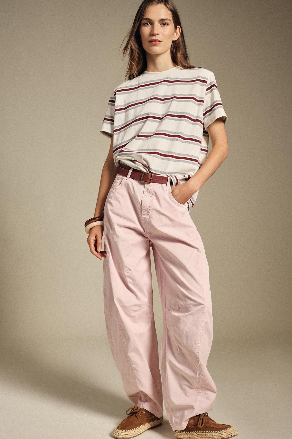 Woman wearing cream striped T-shirt, high-waist pink wide-leg trousers, brown belt and tan suede loafers on neutral studio background