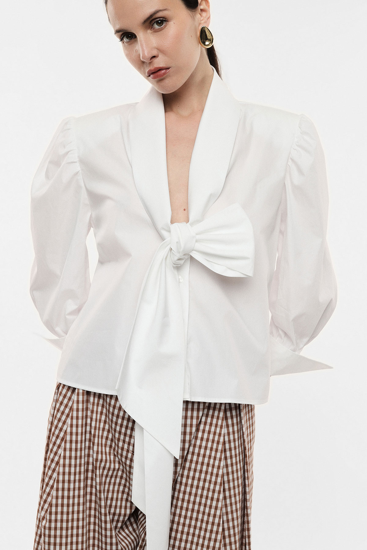 Woman wearing white blouse with oversized front bow and puffed sleeves, gold hoop earring, paired with brown gingham skirt