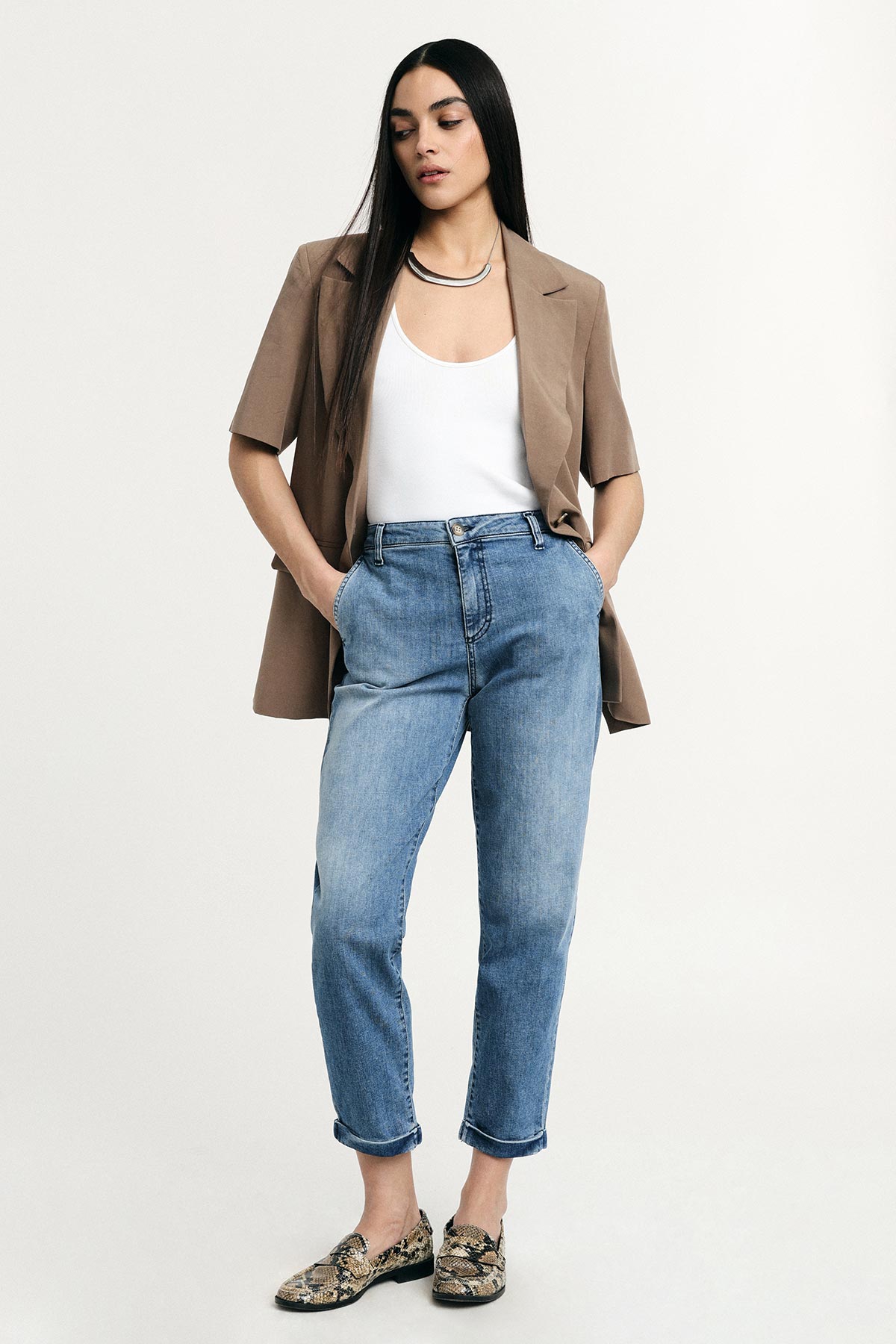 Woman wearing beige short-sleeve blazer, white tank top, high-waist blue jeans and snakeskin loafers, hands in pockets on neutral background