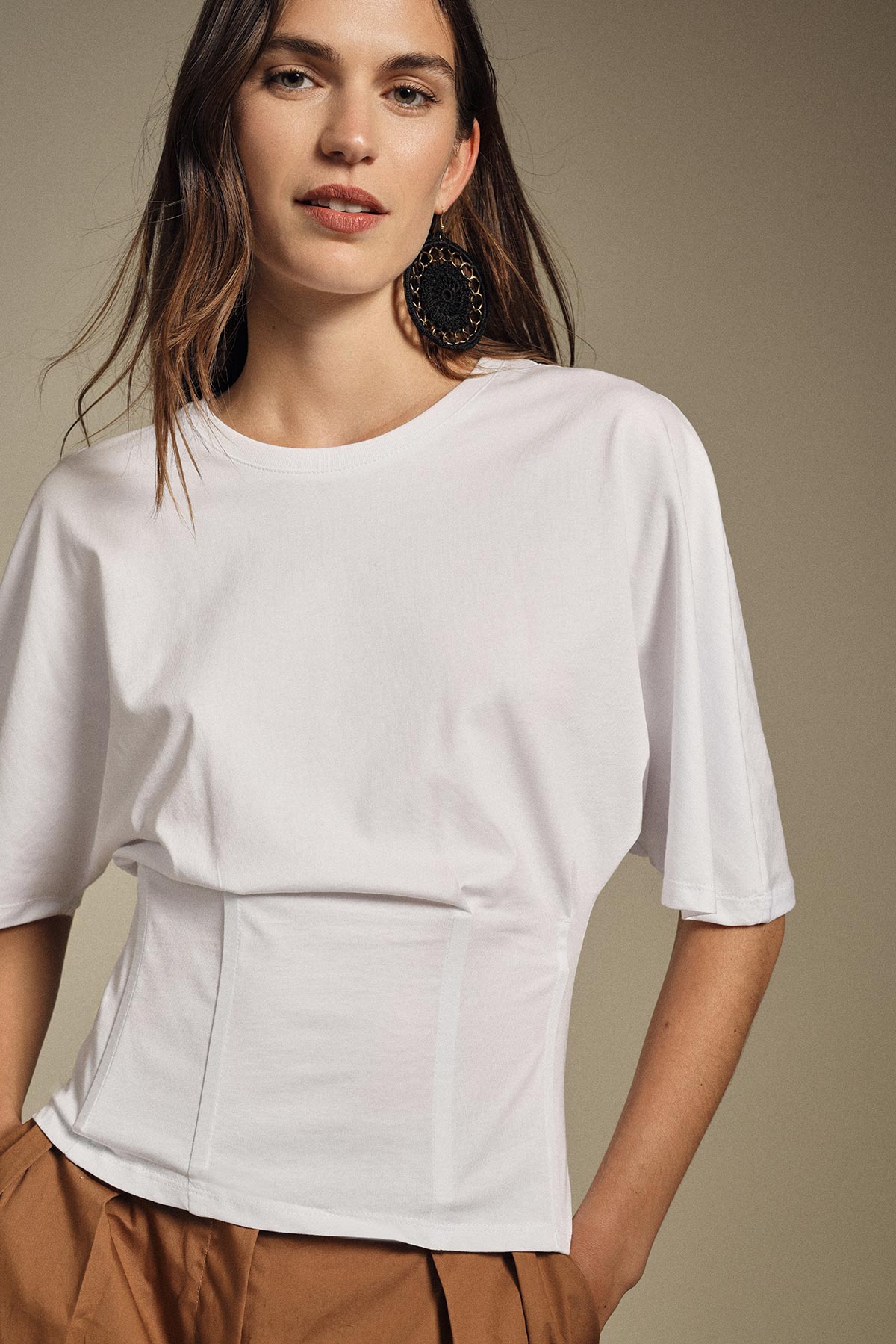 Woman wearing a white short-sleeve fitted top and tan trousers with a large black statement earring against a neutral background