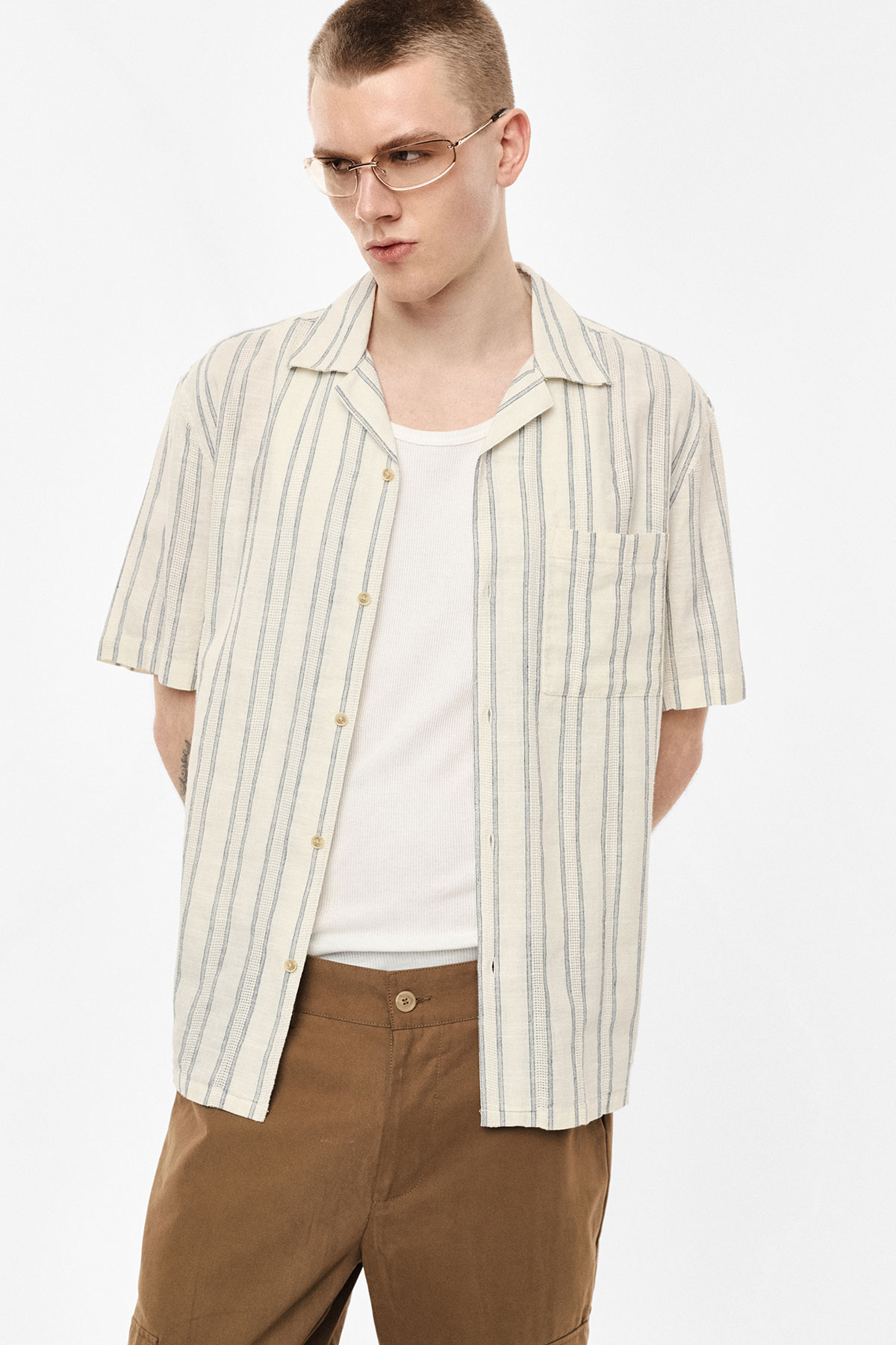 Model wearing cream short-sleeve vertical-striped shirt over white vest, brown trousers and glasses