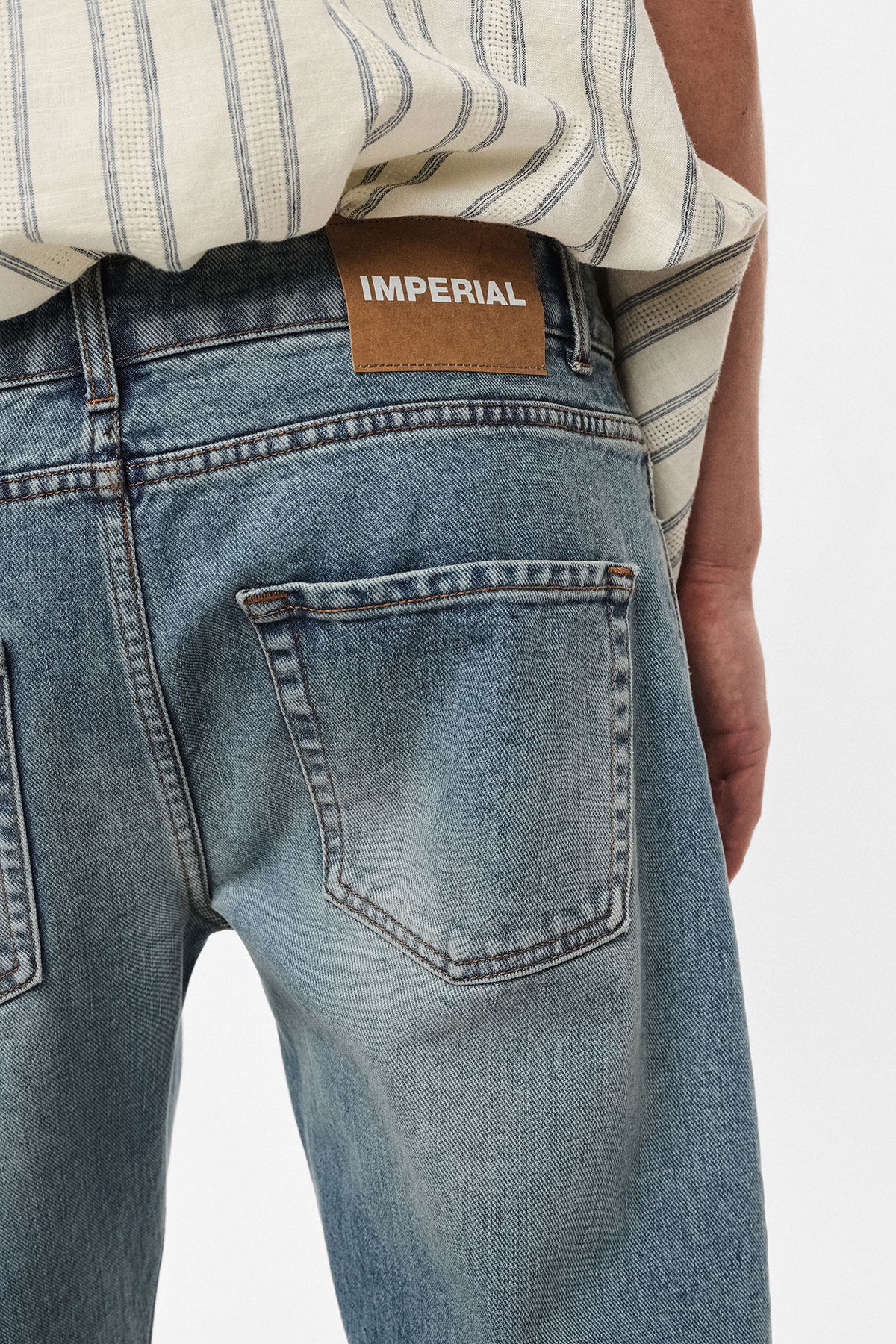 Rear view of faded blue denim jeans with a back pocket and brown leather label reading 'IMPERIAL', striped top visible