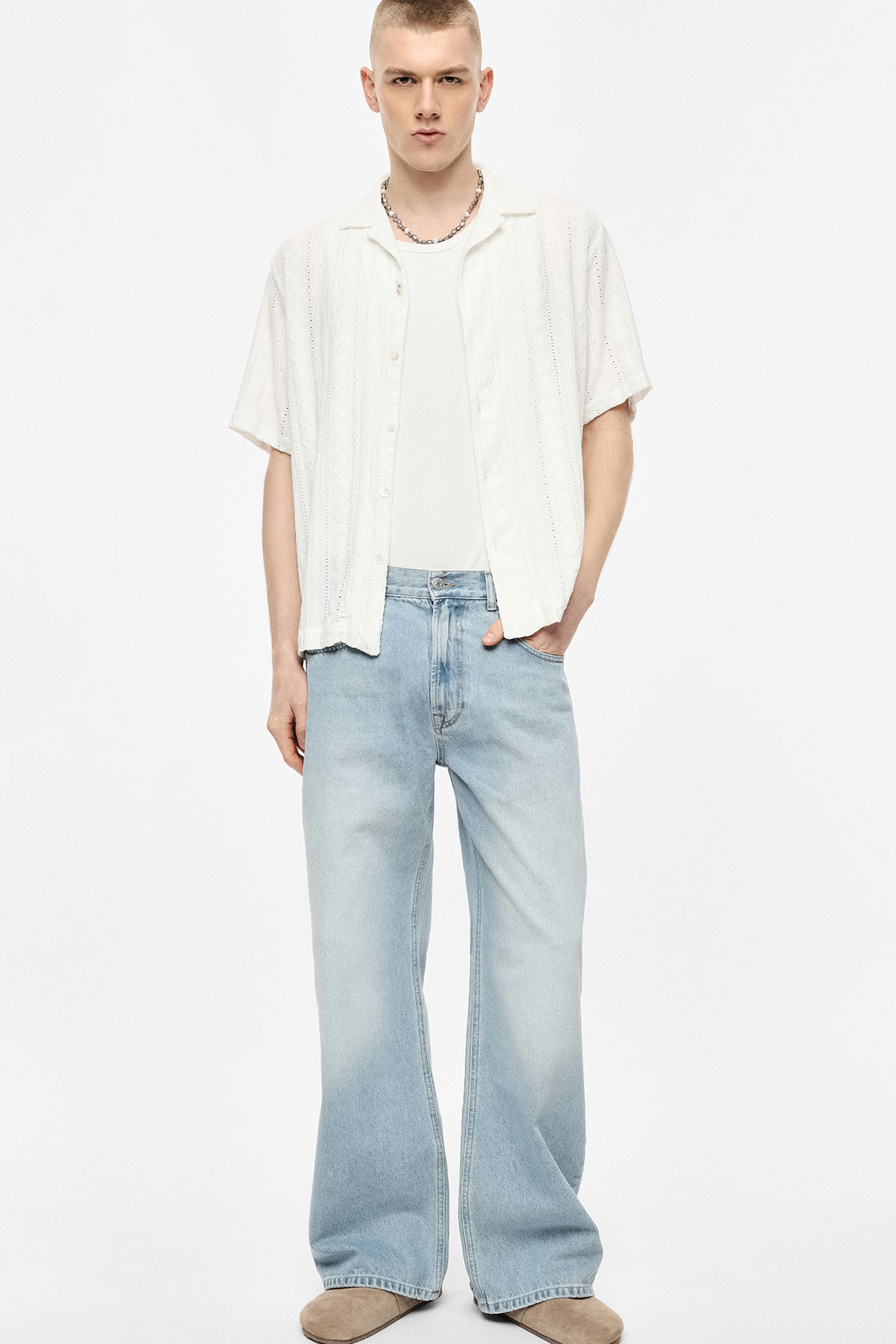 Male model in white open short-sleeve shirt over white vest, chain necklace and light-wash wide-leg jeans, one hand in pocket