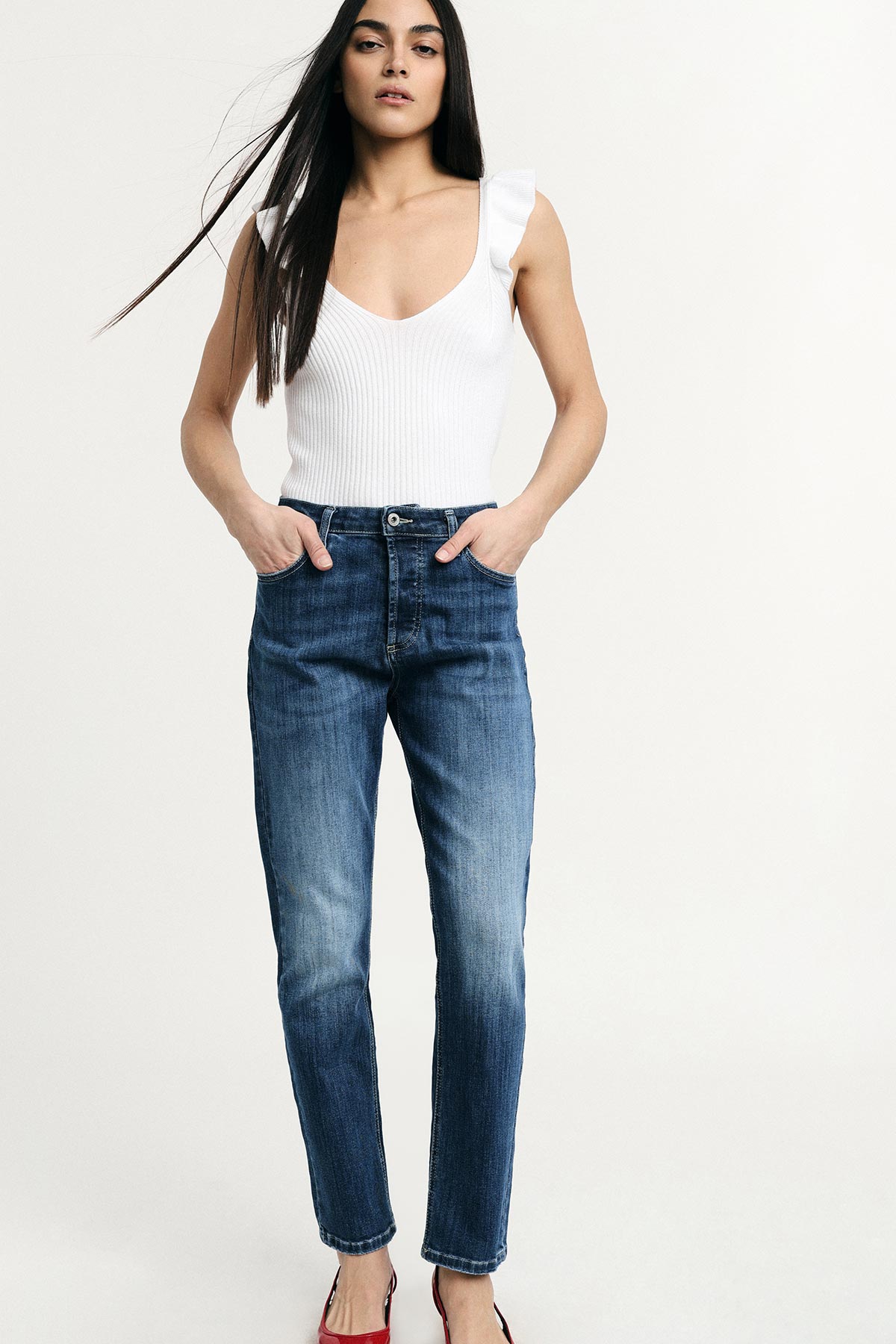 Woman in white sleeveless top and high-waisted blue jeans, hands in pockets, standing against pale background