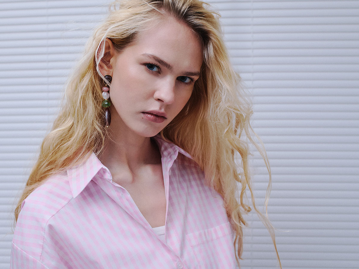 Blonde woman in a pink striped shirt and statement earrings posing against white blinds