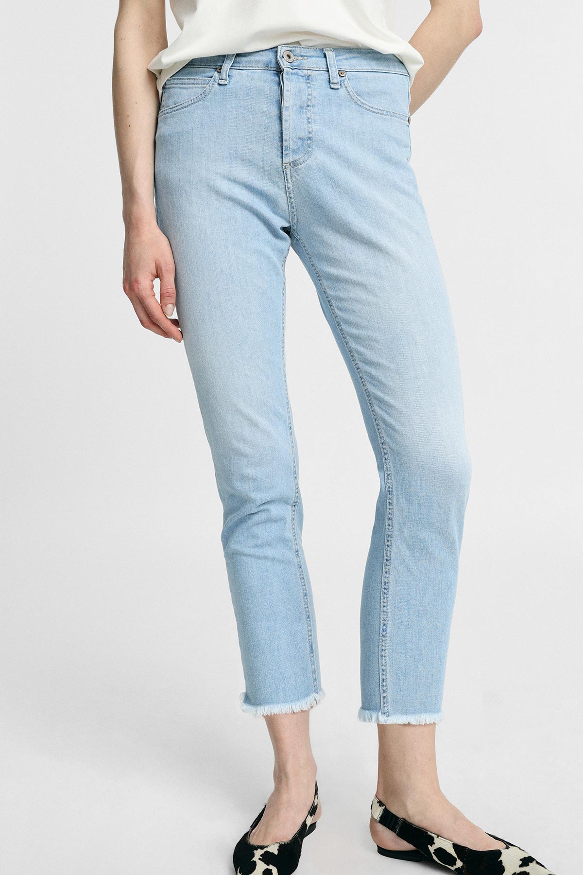 Model wearing light-wash cropped skinny jeans with raw frayed hem, high waist five-pocket style paired with white top and black patterned slingback flats