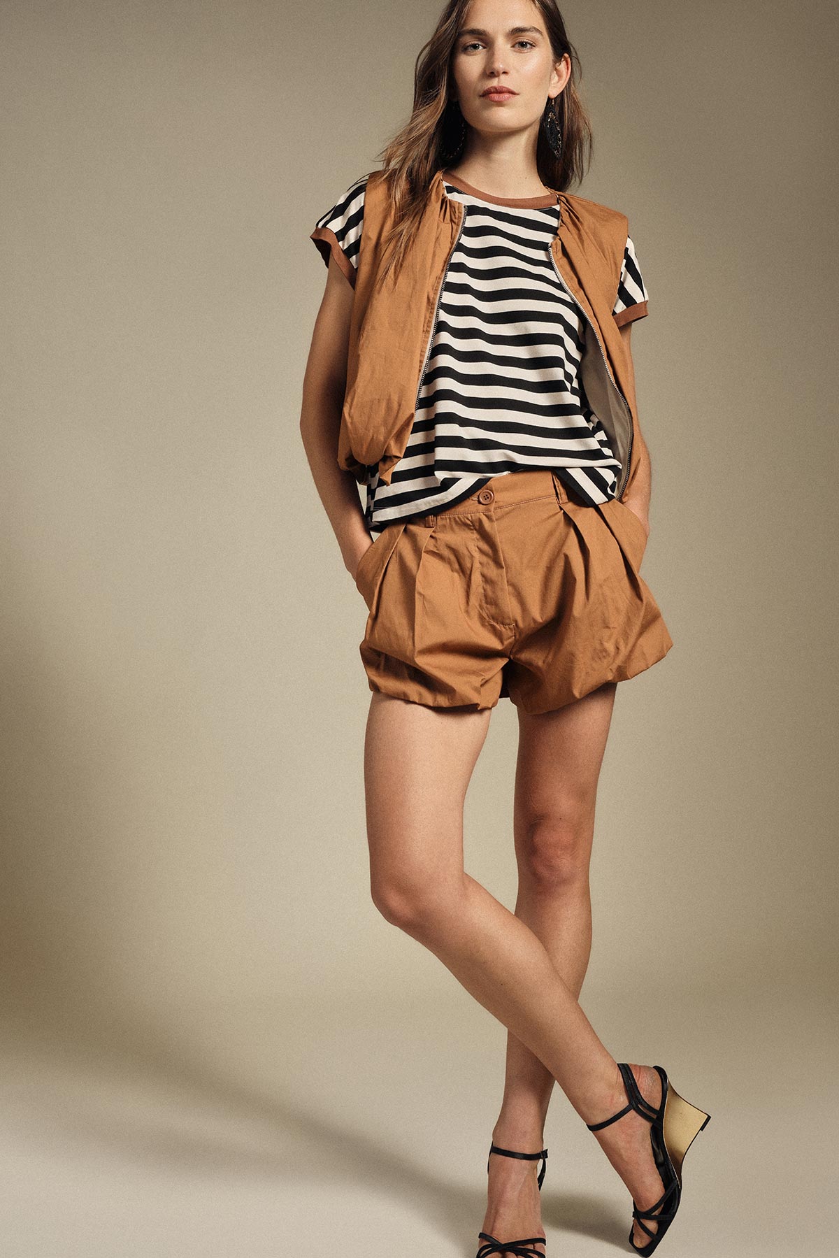 Woman modelling black-and-white striped t-shirt, tan sleeveless jacket and matching pleated shorts with black strappy heels, standing pose