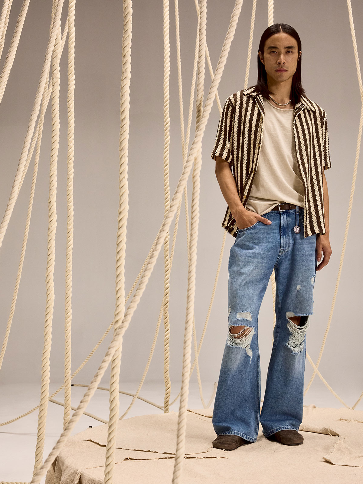 Young person in striped short-sleeve shirt, beige tee and ripped wide-leg jeans standing amid hanging ropes on a beige studio set