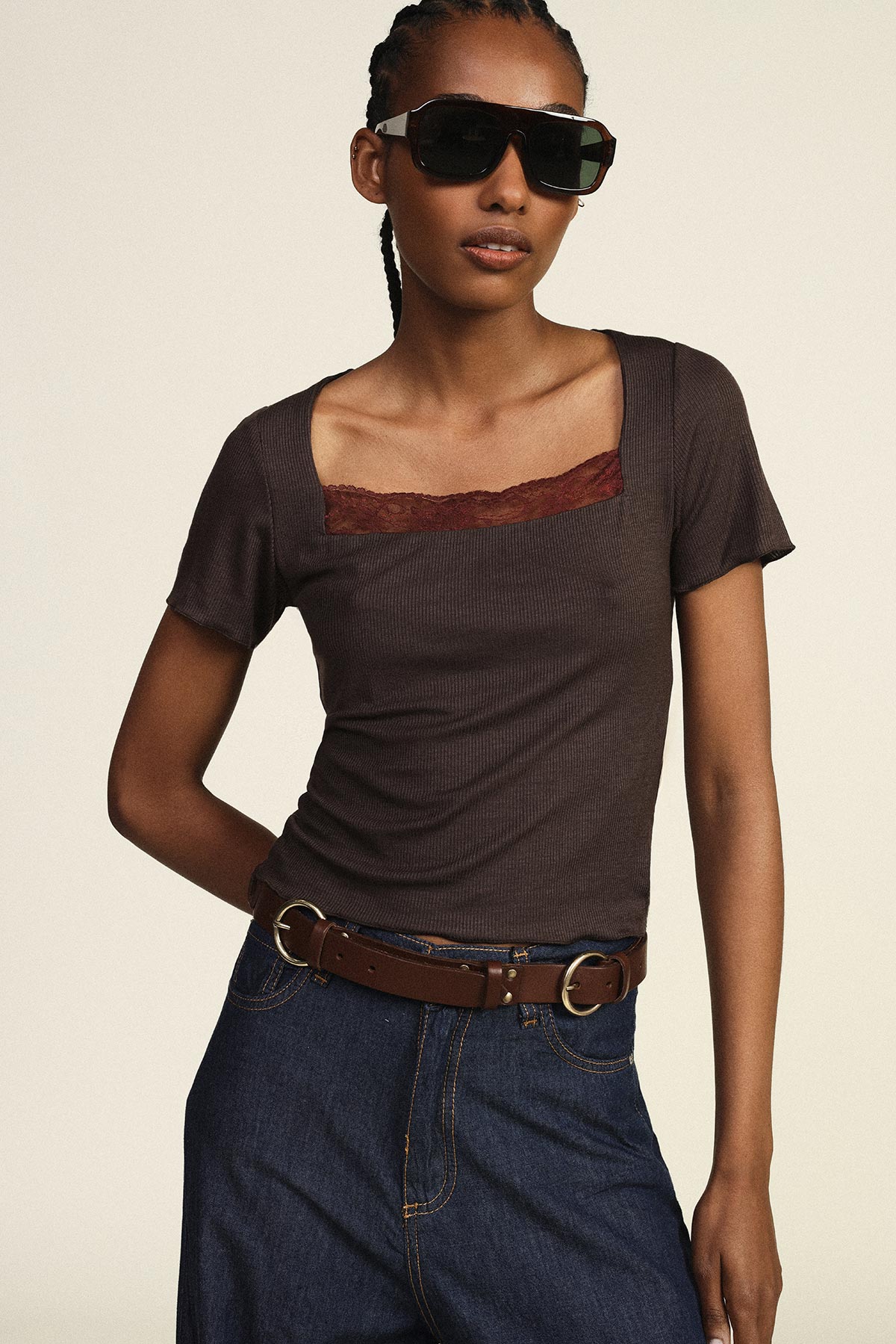 Woman modelling dark square‑neck ribbed top with lace trim, denim skirt, brown belt and oversized sunglasses on beige background