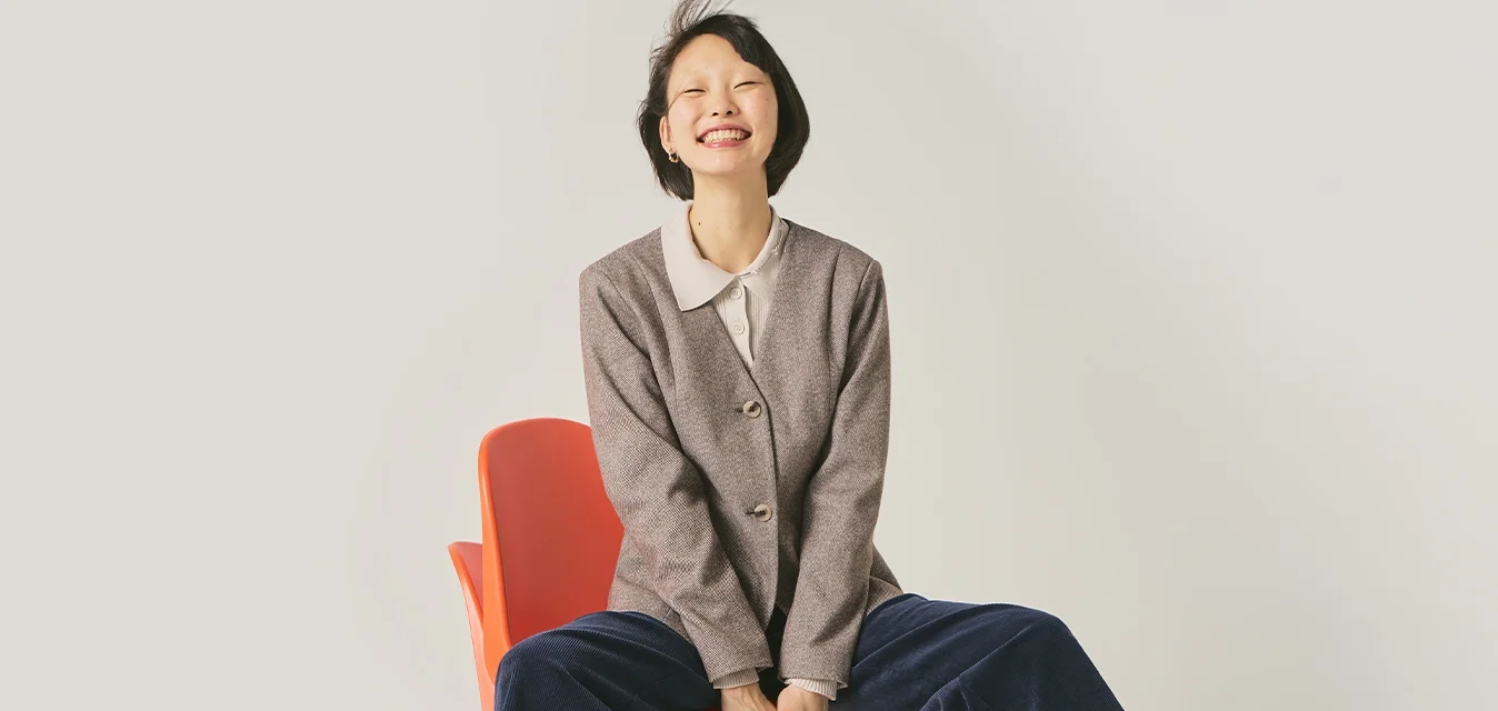 Giovane donna sorridente, seduta su una sedia arancione con le gambe divaricate, indossa un blazer grigio, pantaloni larghi blu navY e stivali scuri su uno sfondo semplice.