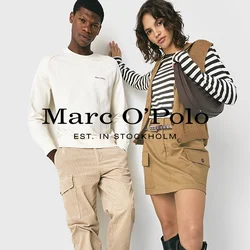 Due adulti appoggiati a un muro bianco, uomo in felpa e pantaloni color crema, donna in top a strisce, gonne beige, gilet, stivali neri, che porta una borsa.