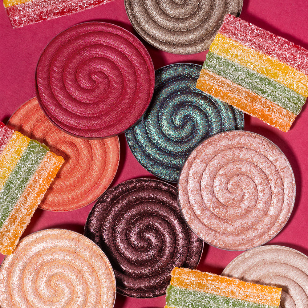 Close-up of colourful shimmery spiral eyeshadow pans and sugared rainbow gummy slices on a pink background