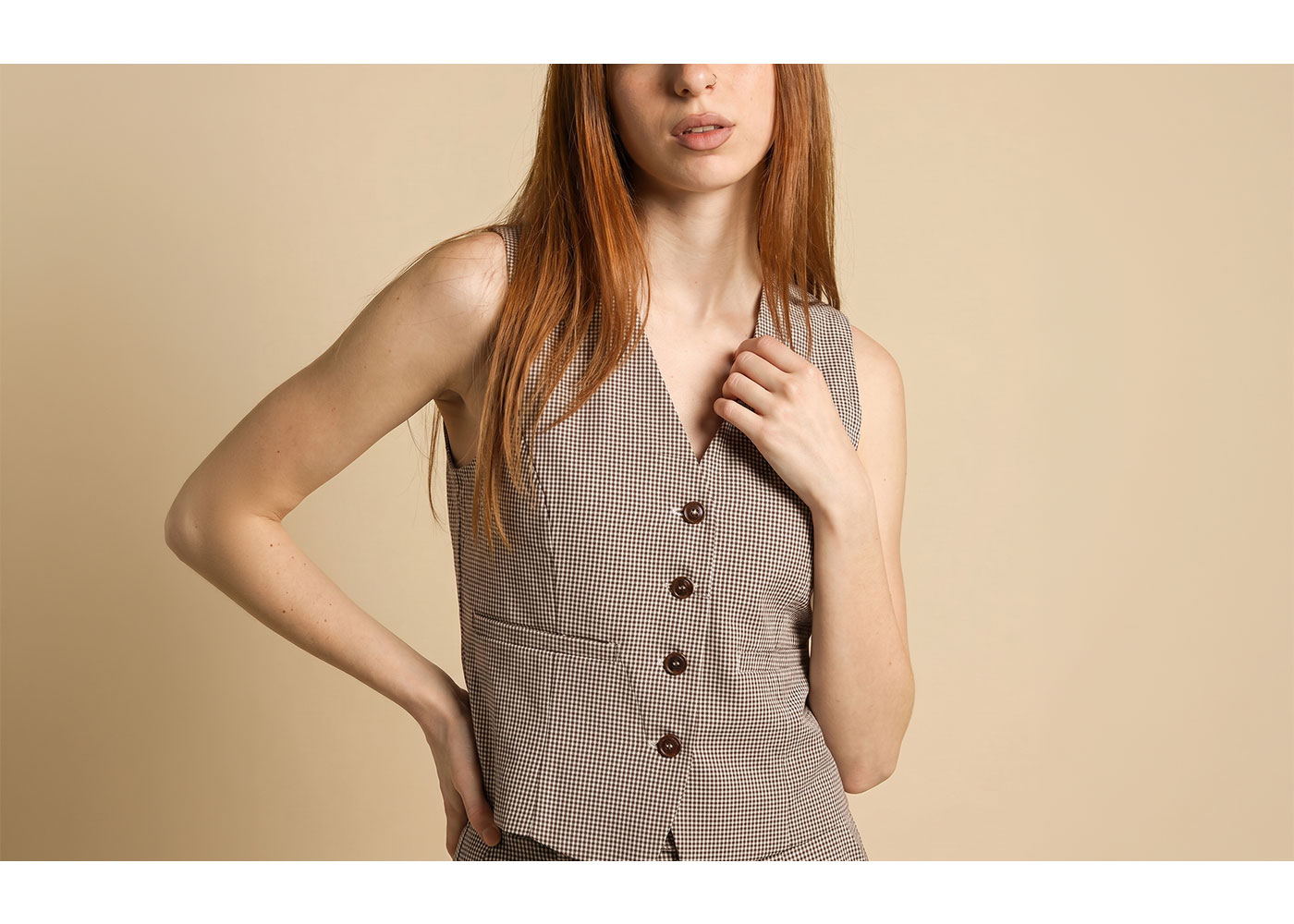 Red-haired woman modelling brown checked waistcoat against beige background, cropped torso with hand on chest and hip