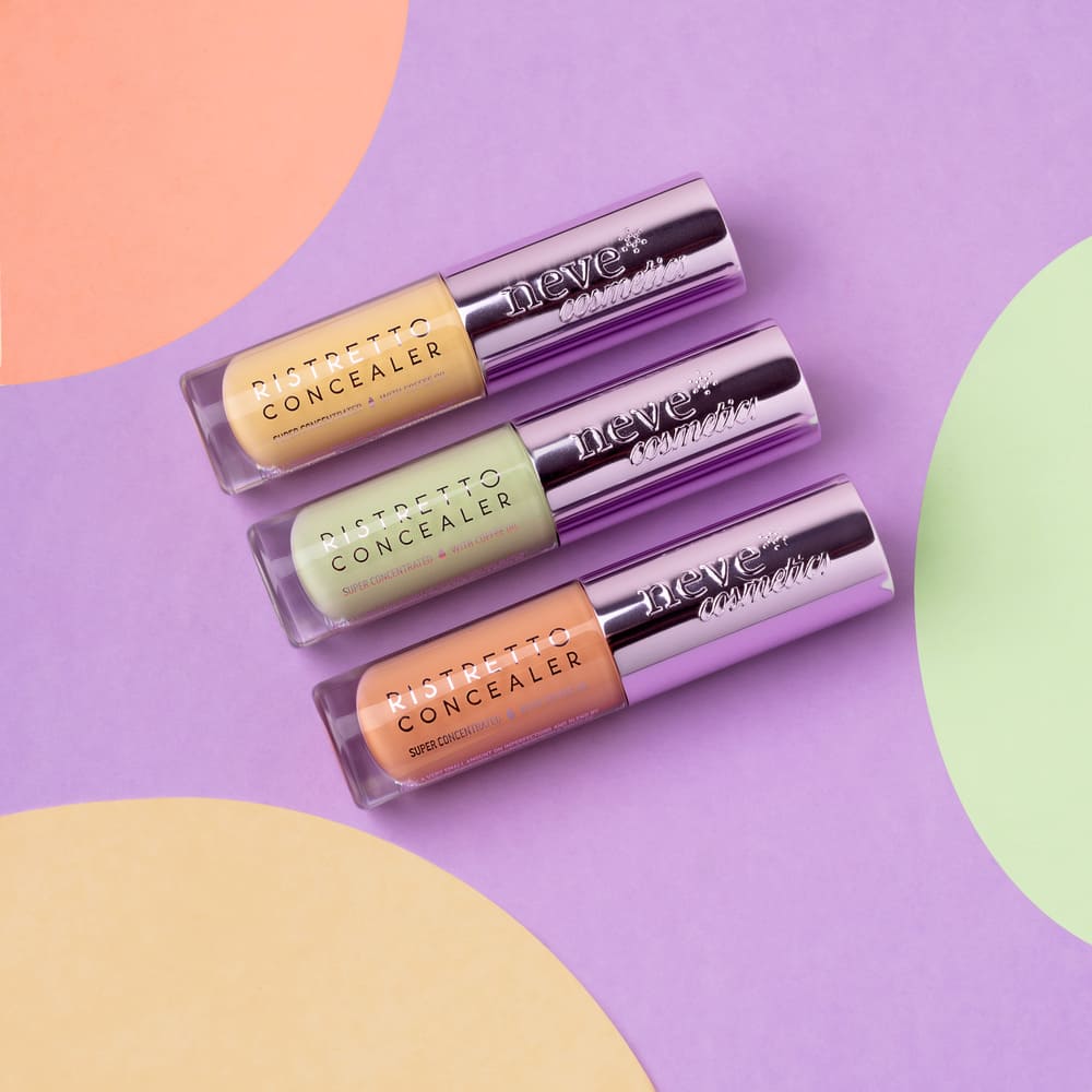 Three Neve Cosmetics Ristretto concealer tubes in yellow, green and peach on a purple background with pastel circular accents