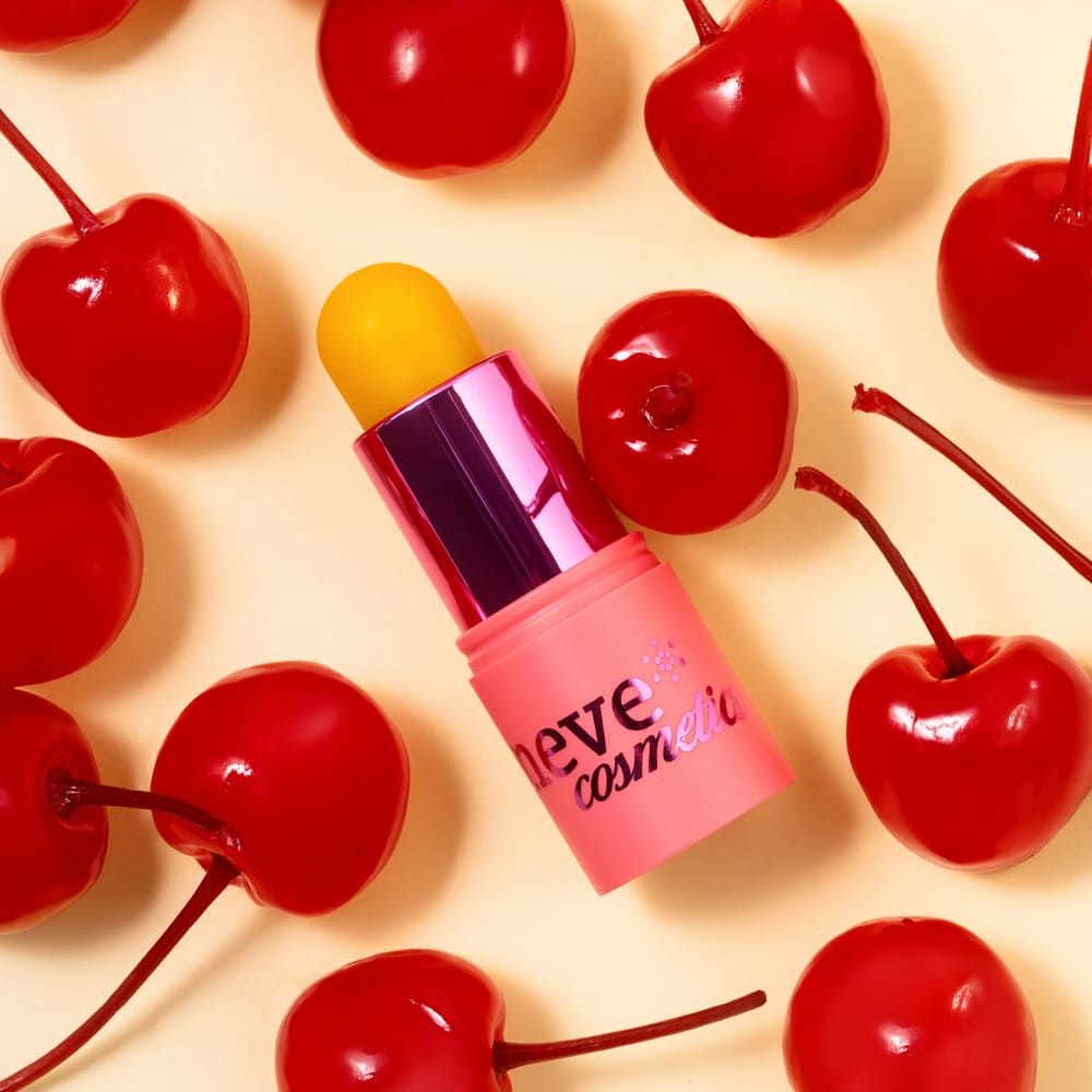 Yellow lip balm in a pink 'Neve Cosmetics' tube surrounded by glossy red cherries on a pale background