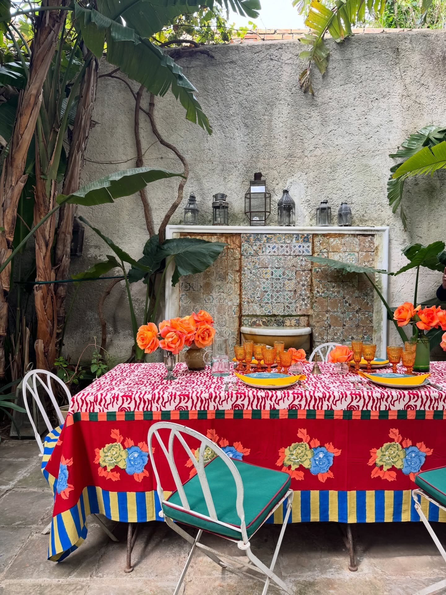 Outdoor dining table with patterned tablecloth, orange roses, amber glassware, tiled fountain and tropical plants