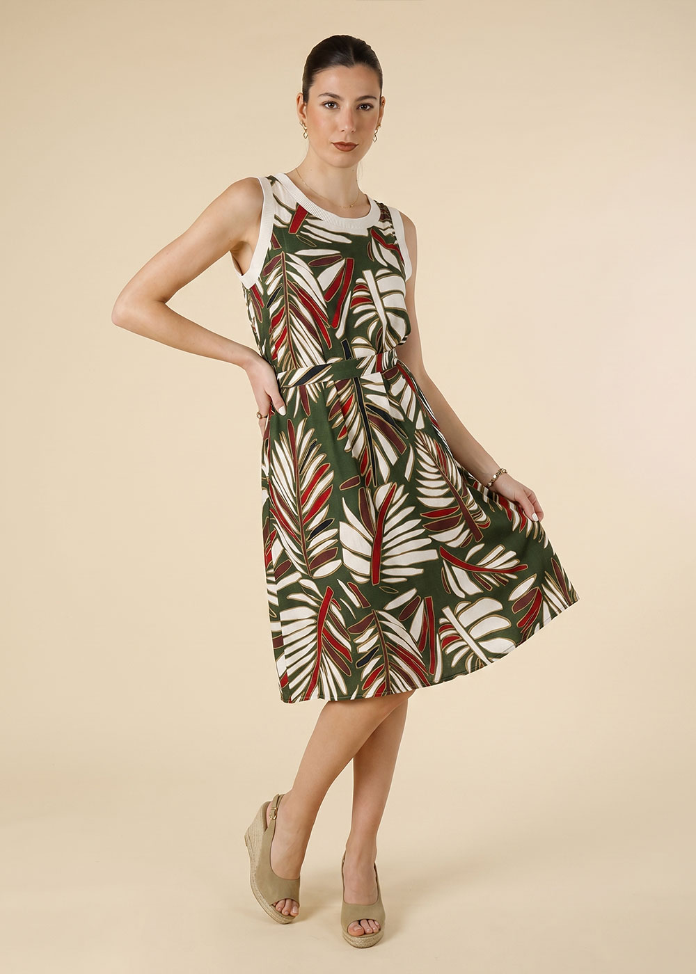 Woman modelling green tropical leaf-print sleeveless knee-length summer dress with waist tie and beige wedge sandals on neutral background