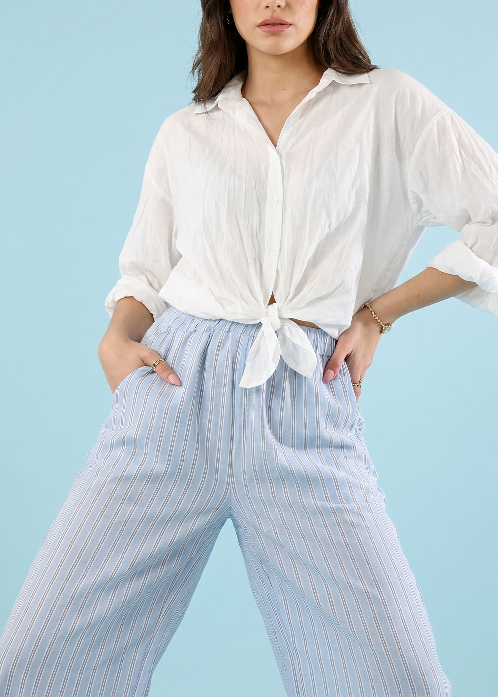 Model wearing tied white blouse and light blue vertical striped wide-leg trousers, hands in pockets on pale blue background