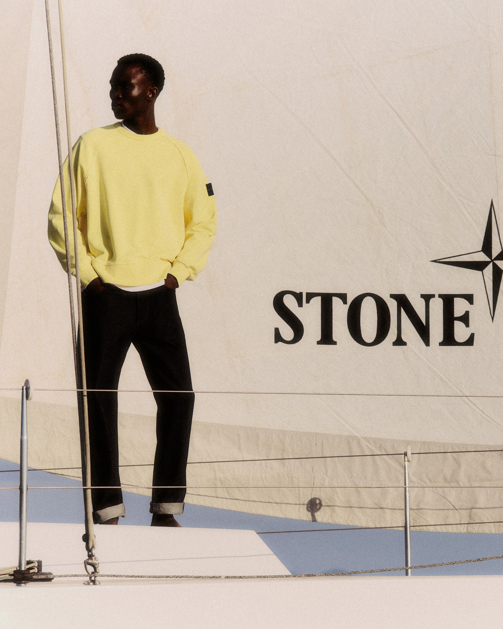 Person standing on yacht deck wearing yellow sweatshirt and dark trousers beside a sail branded 'STONE'