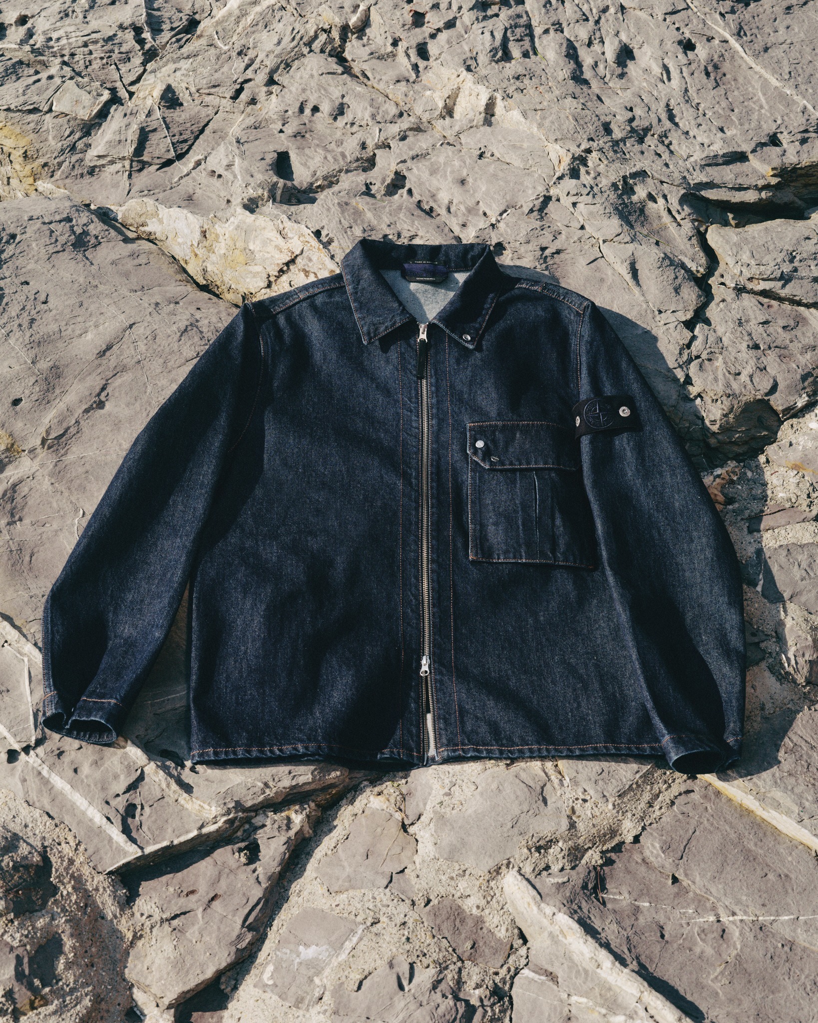 Dark denim zip-front jacket with chest pocket laid flat on textured rocky surface — men's casual outerwear flatlay
