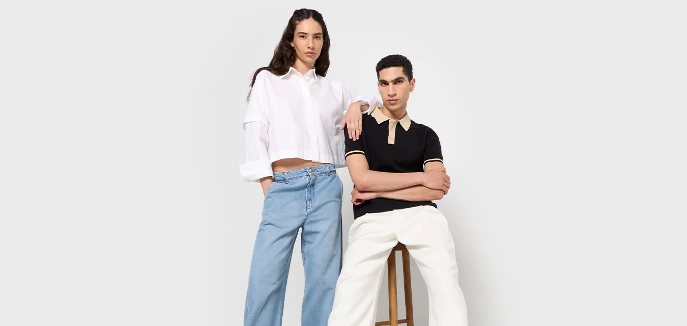 Due modelli stanno uno accanto all'altro. Uno indossa una maglietta corta bianca e jeans ampi blu; l'altro indossa un polo nera e pantaloni bianchi.
