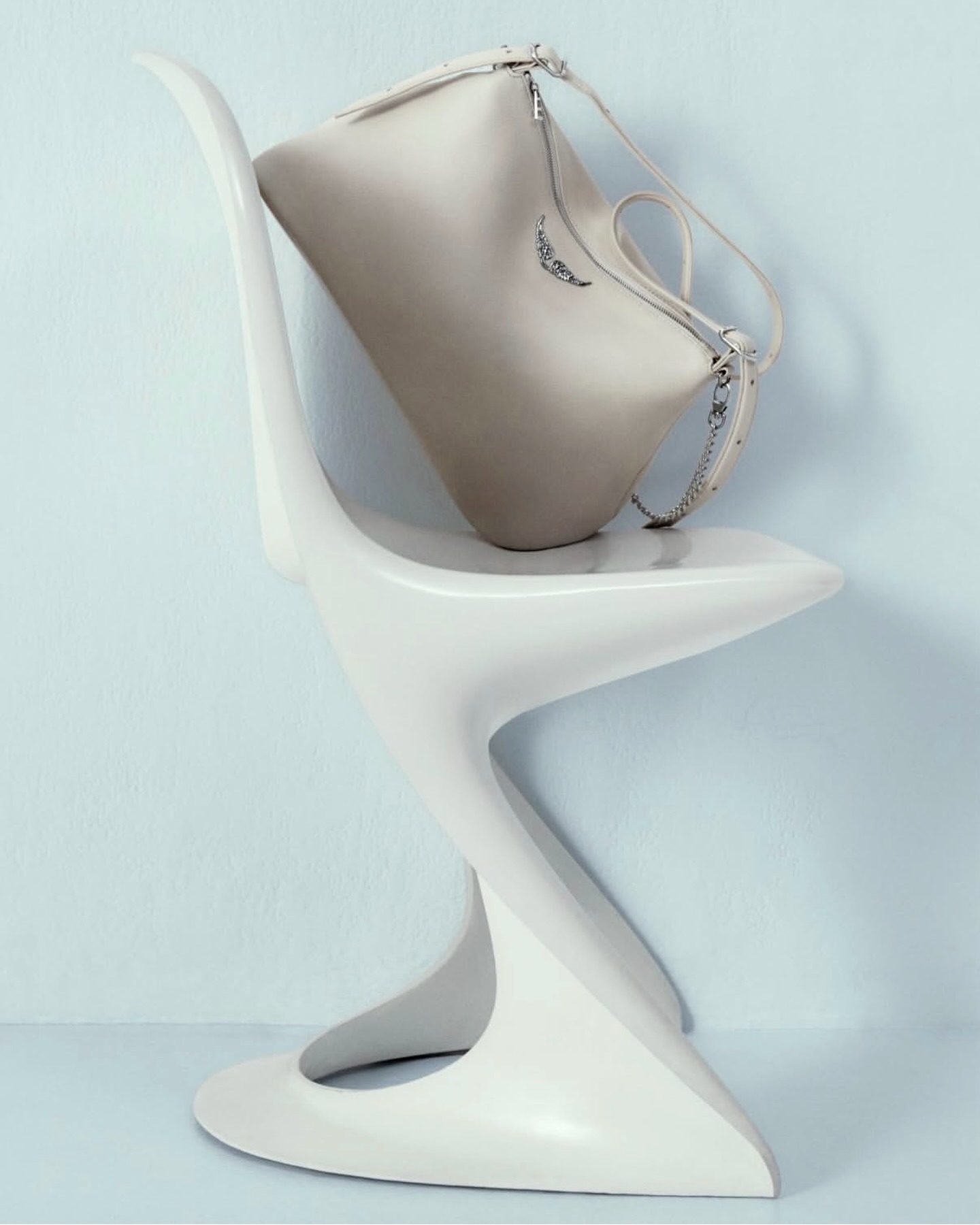 Beige leather handbag with silver chain strap resting on a modern white sculptural chair against a pale blue wall