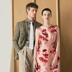 Uomo con blazer verde oliva e pantaloni beige sta accanto a una donna con un vestito floreale rosa su uno sfondo beige minimalista.