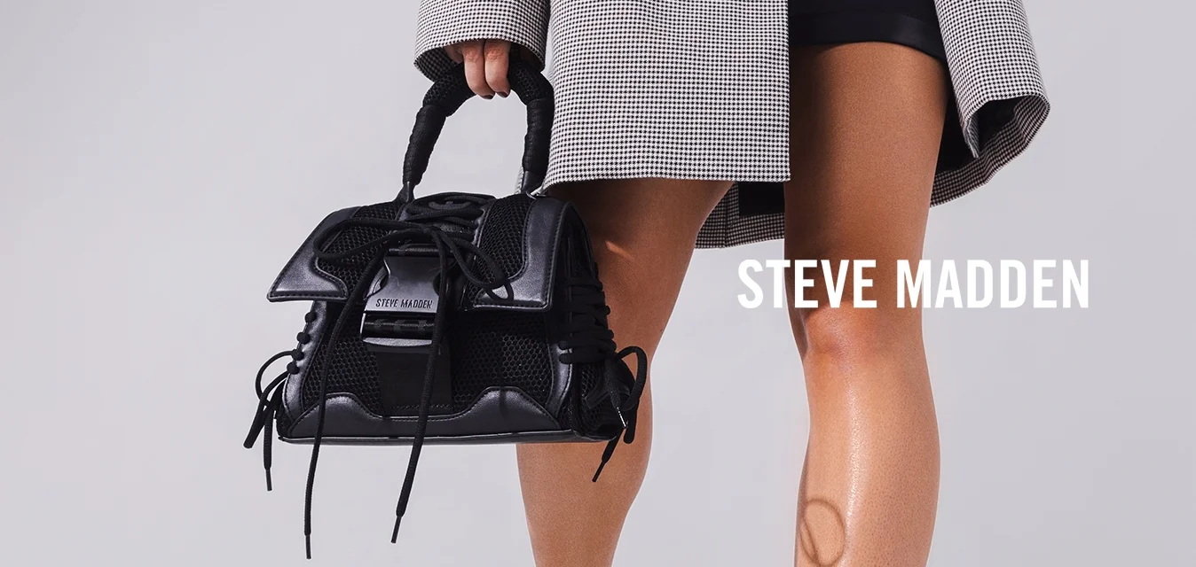 Donna che indossa un blazer a quadri oversize sopra un crop top nero e pantaloncini, tiene una borsa nera in mano, in piedi in una stanza bianca con lucernario.