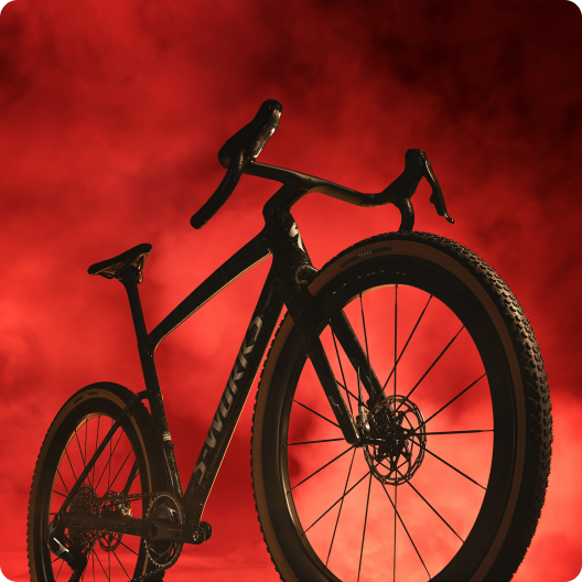 Black gravel bike with drop handlebars and disc brakes photographed against a dramatic red smoky studio backdrop
