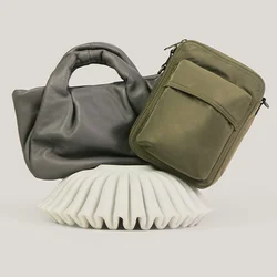Due borse, una borsa tote in morbida pelle grigia e una tracolla rettangolare verde oliva, esposte su un supporto bianco testurizzato.